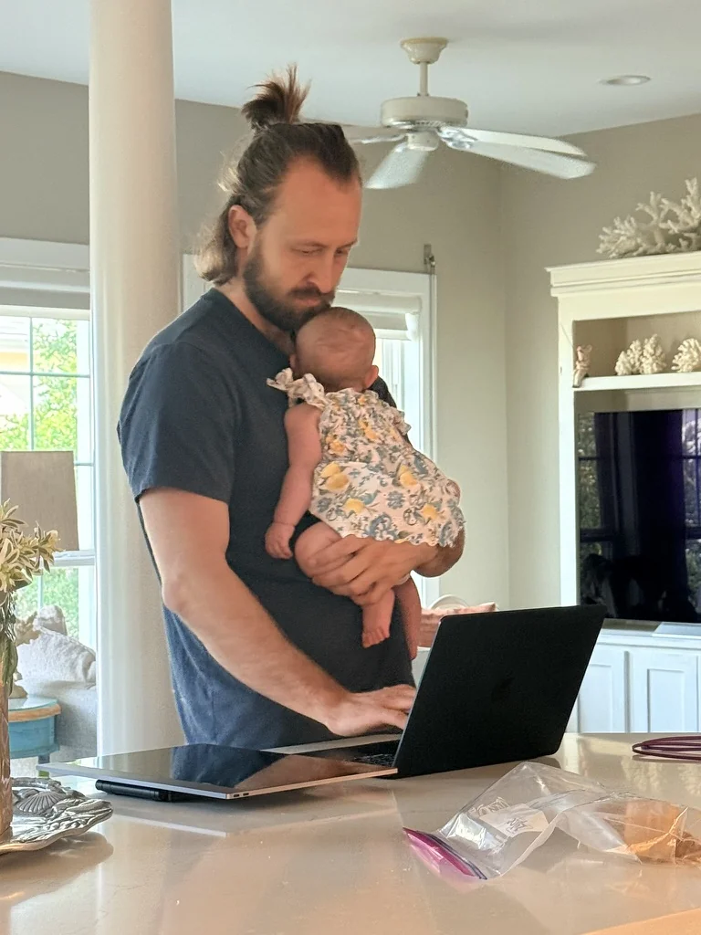 Andrew holding his infant daughter while working at his laptop